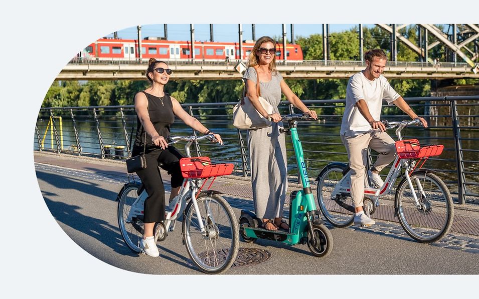 2 Personen auf Fahrrad und 1 Person auf Scooter freudig fahrend und im Hintergrund Brücke mit einer Eisenbahn