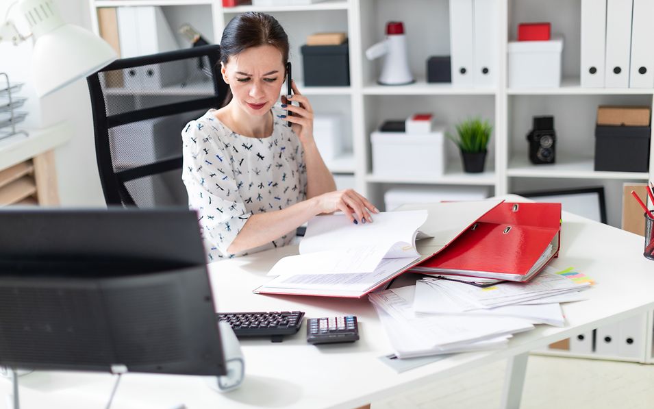 Frau sitzt am Schreibtisch vor aufgeschlagenen Aktenordnern und einem PC Bildschrim und prüft Unterlagen